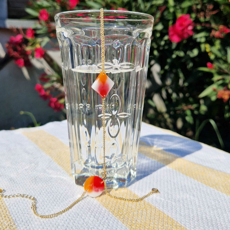 Bracelets l'Iconique rond et carré posés sur un verre transparent. L'Iconique est un bracelet composé d'un rond ou d'un carré de résine colorée (ici, la gamme Lantana Camara: Rouge, orange et blanc) et d'une fine chaine plaquée or.