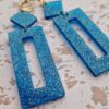 Boucles d'oreilles (fermoirs dormeuses plaqué or 24k) en résine bleue à paillettes composée d'un petit carré et d'un rectangle plus long.